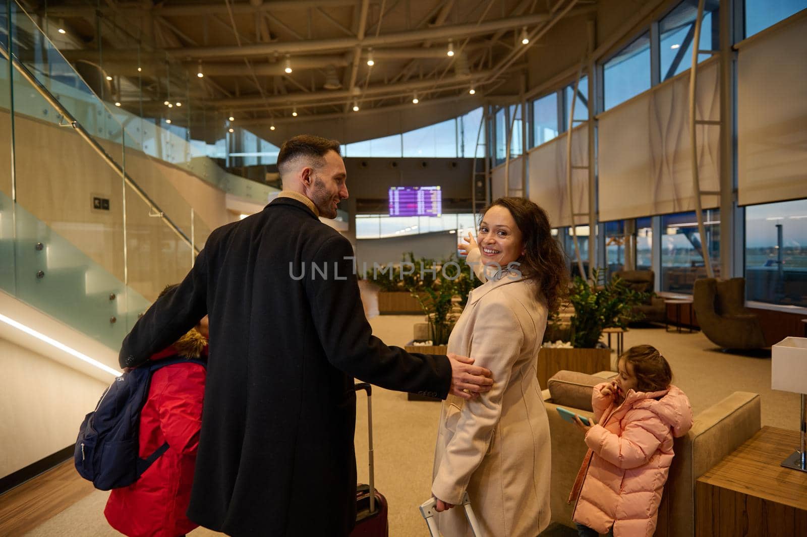 Loving father hugs his son and wife as she smiles, looking over her shoulder, pointing to timetable information panel, checking the flight at the airport departure terminal. Family air travel concept