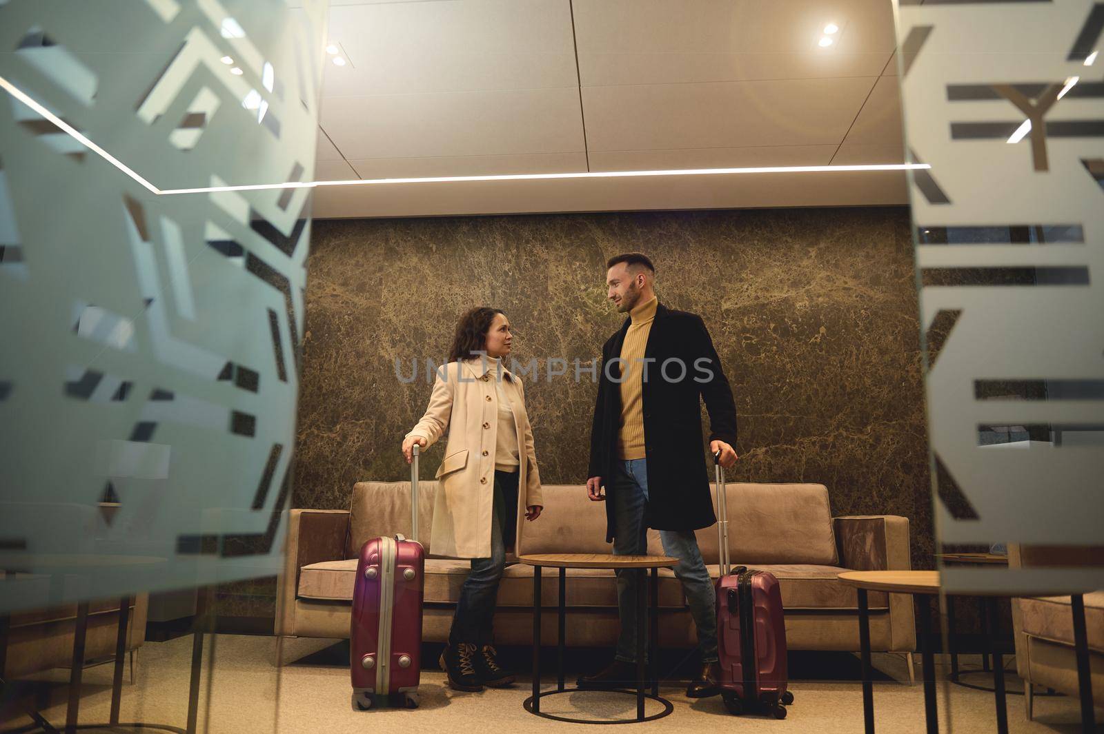 Handsome attractive businessman talking to his business partner, beautiful woman standing with suitcases at the exit from the conference room in the airport terminal ready to board the flight together