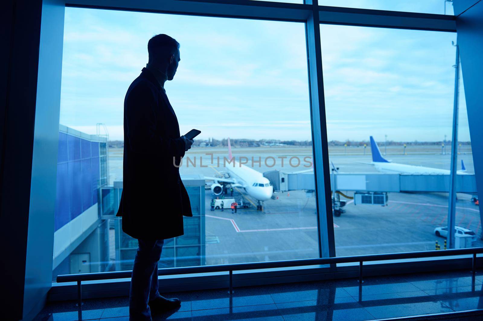 Silhouette of businessman in airport lounge with mobile phone standing by panoramic windows overlooking runways and airplanes waiting for flight early in the morning. Business trip, air travel concept