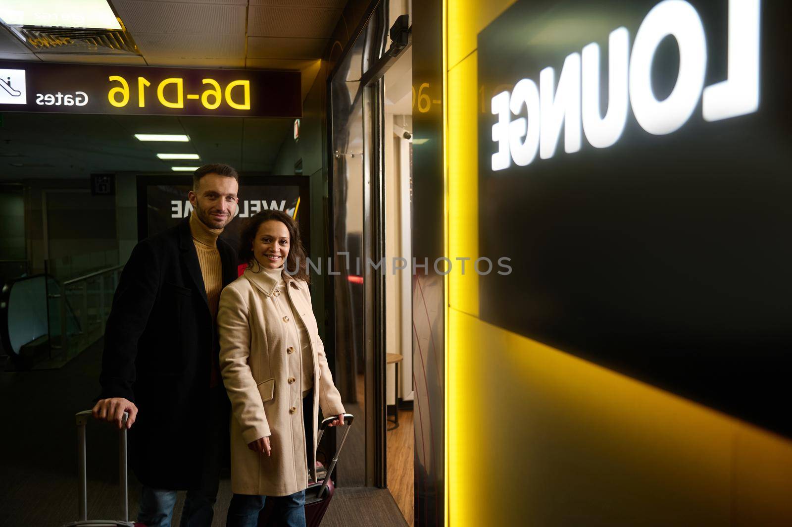 Business partners, multiracial couple of a middle aged woman and man with suitcases smiling looking at camera at the entrance to the duty-free area of the departure terminal of international airport by artgf