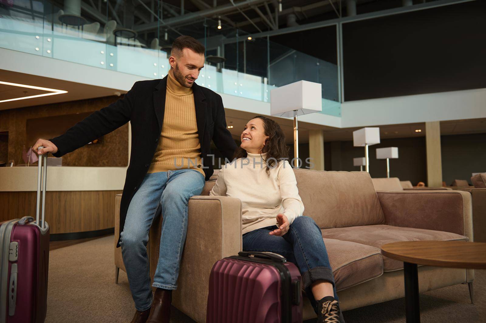 Woman and man in casual clothes with suitcases sitting on armchair in the international airport terminal lounge, looking at each other, discussing forthcoming flight, waiting for airplane departure by artgf