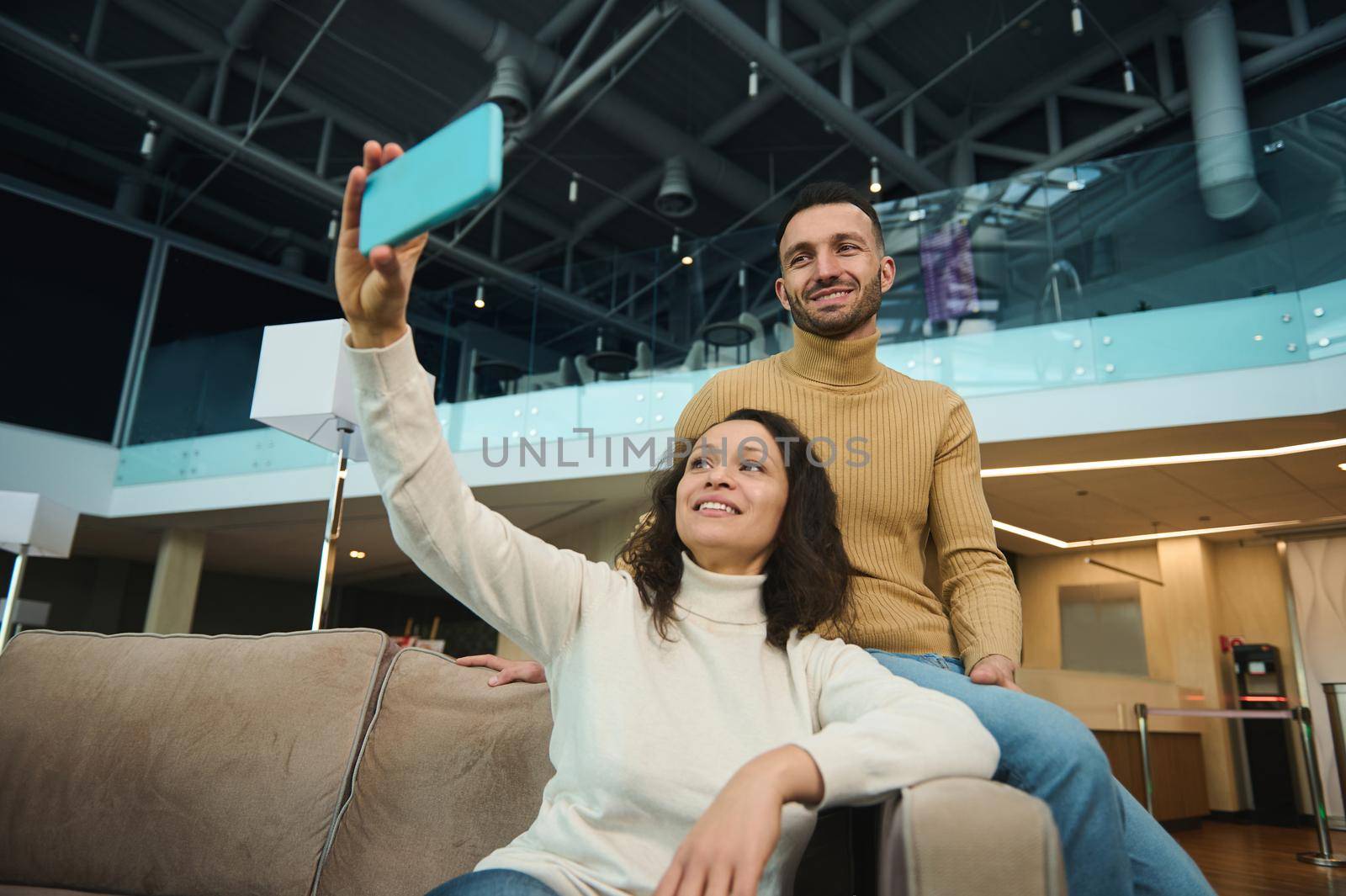 Beautiful young loving couple of newlyweds on their honeymoon trip making a self-portrait selfie on a smartphone while relaxing together in the airport lounge while waiting for boarding a flight.