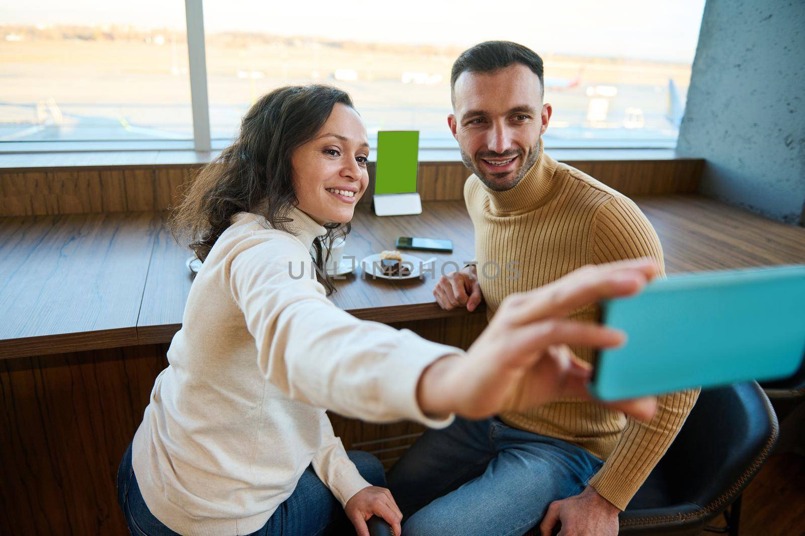 Beautiful multi ethnic loving couple enjoying coffee break together, sitting at a table in an airport restaurant, taking a selfie against the backdrop of runways with planes, waiting to board a flight