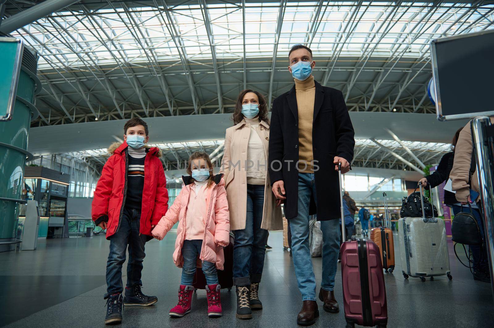 Multiracial young happy family at the airport with luggage while waiting for a flight in a pandemic. Winter air travel concept.