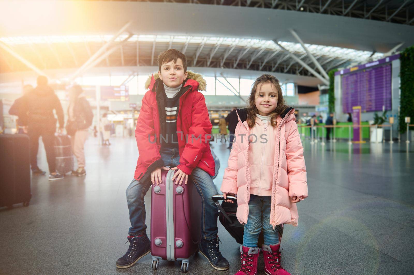 Happy beautiful Caucasian children in warm clothes, a handsome boy sits on a suitcase next to his younger sister- a cute little girl, at the international airport. Family airplane travel concept