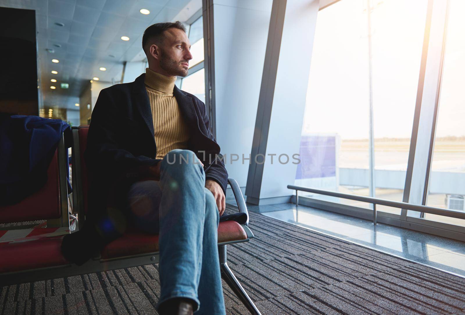 Handsome middle aged confident businessman on business trip, sitting in the departure lounge, admiring the view from the panoramic windows of the international airport to the runway with airplanes