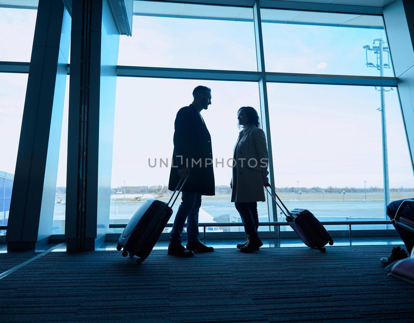 Silhouette of two people, woman and man with suitcases, standing by the panoramic windows overlooking the runways and planes in the departure terminal of the international airport, awaiting a flight by artgf