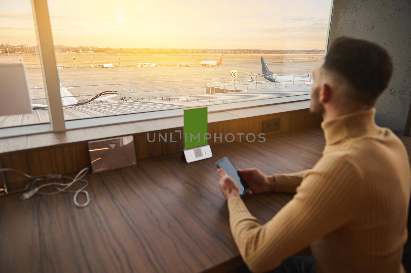 Rear view. Businessman entrepreneur handsome man with smartphone sitting at table by panoramic windows with view of airplanes on runway at sunset, rests in airport lounge cafe while waiting for flight by artgf