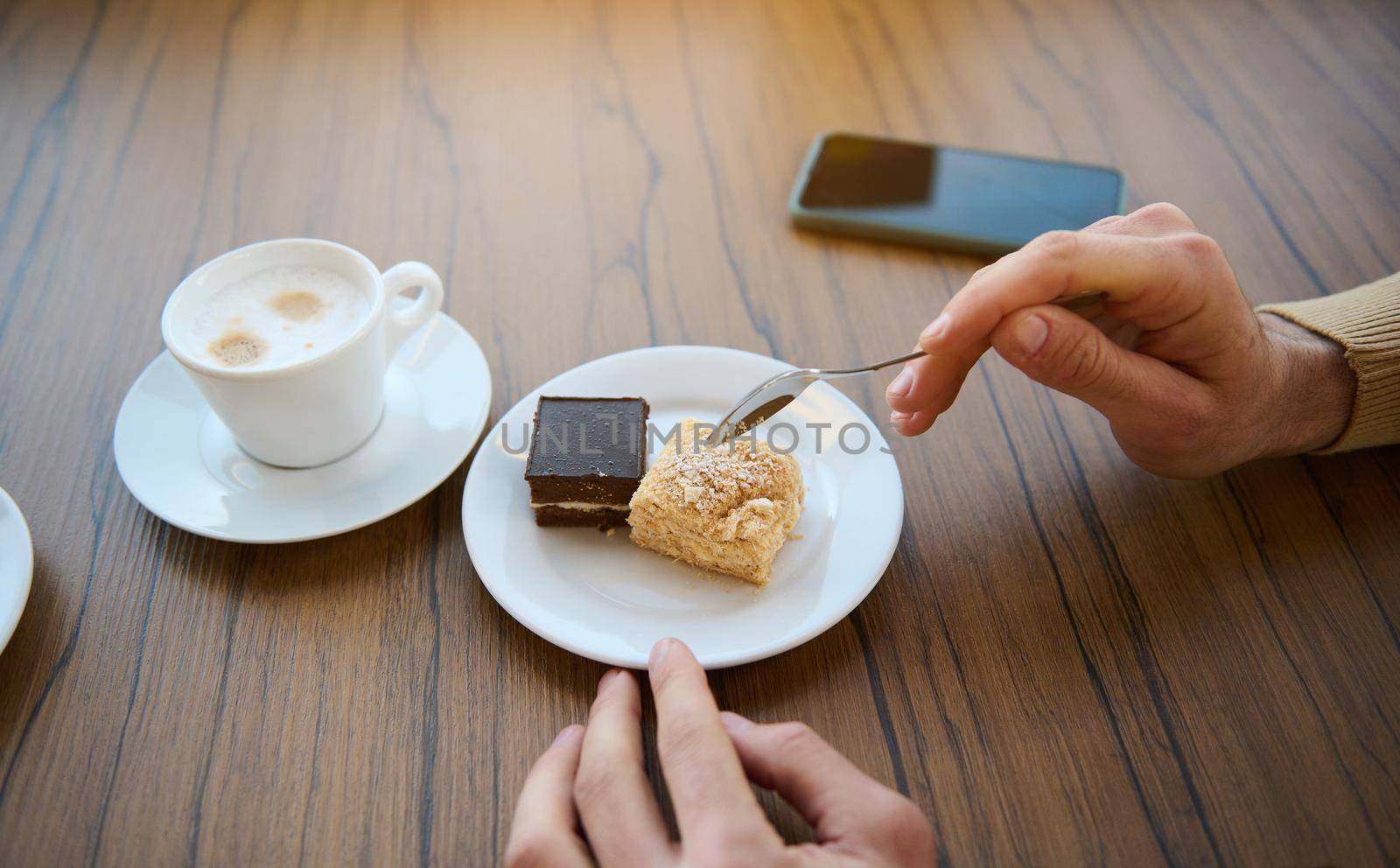 Cropped image of male hands holding tea spoon near delicious sweet dessert, chocolate and cheese creamy cake, cup of aroma coffee with foam and smartphone on wooden table. Coffee break, snack concept by artgf