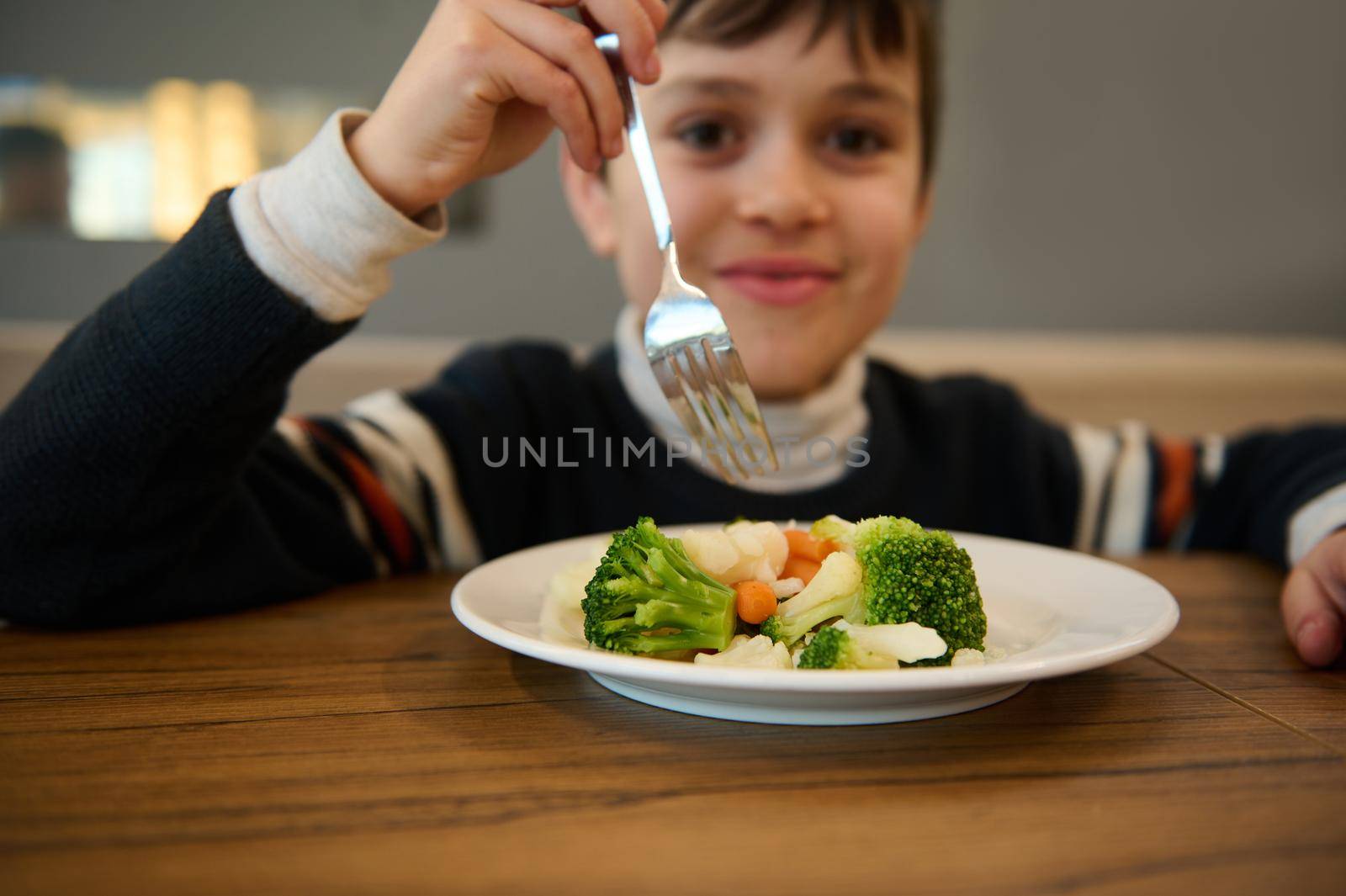 Image focused on a plate of steamed vegetables. Adorable smiling Caucasian teenage boy sitting at table and eating healthy lunch of steamed carrot and broccoli. Nutrition, vegetarian diet for toddler by artgf