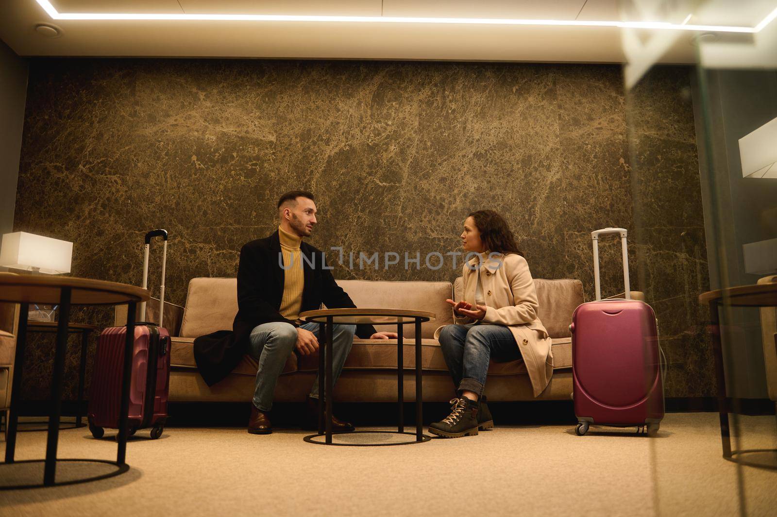 Middle-aged handsome man and pretty woman with suitcases, business partners on a business trip having a conversation in a meeting room while waiting for flight in the airport departure terminal by artgf