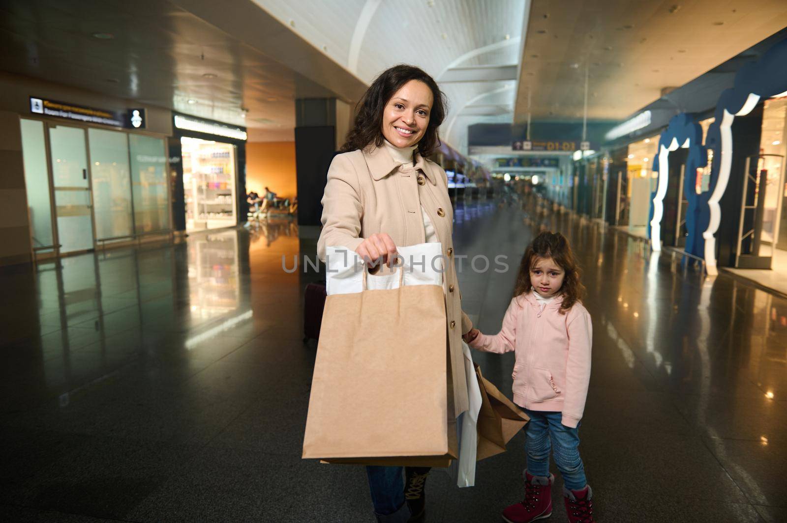 Beautiful woman, loving mother with shopping bags stands with her adorable little daughter in the duty free zone of the departure terminal at the international airport, waiting for the upcoming flight