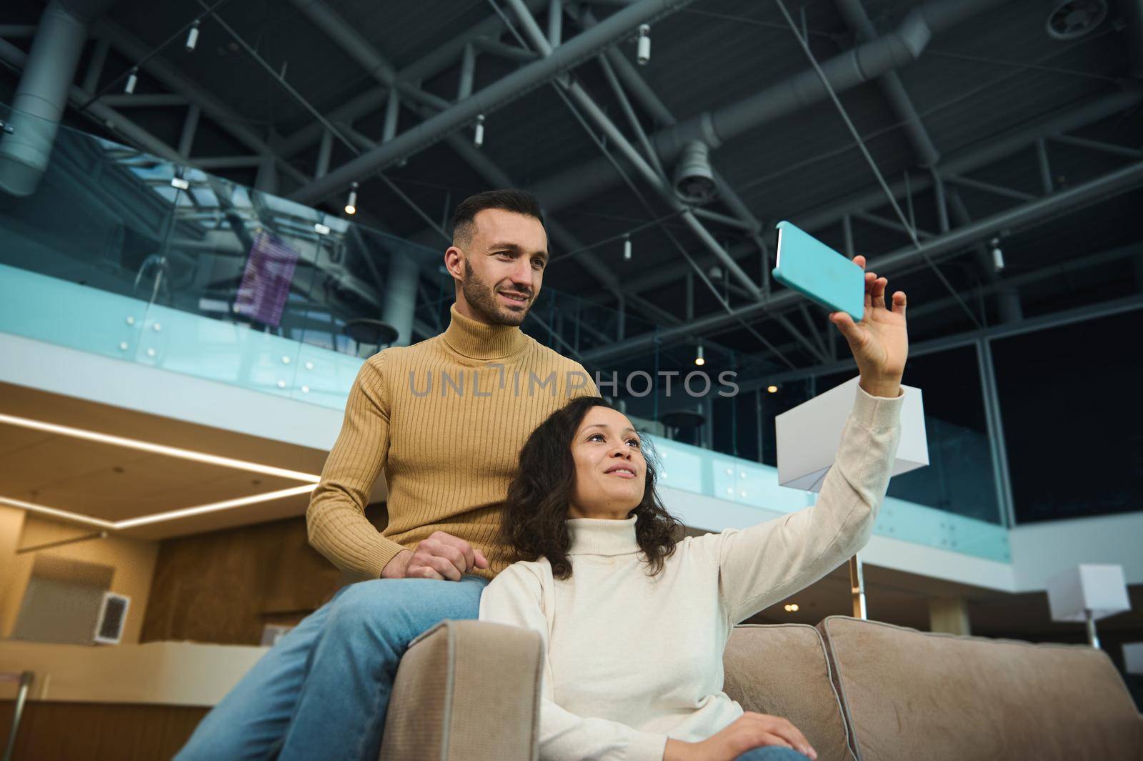 Charming woman and handsome man, friends, newlyweds, multiracial heterosexual couple in love taking selfie while waiting to board a flight in an international airport lounge during honeymoon trip.