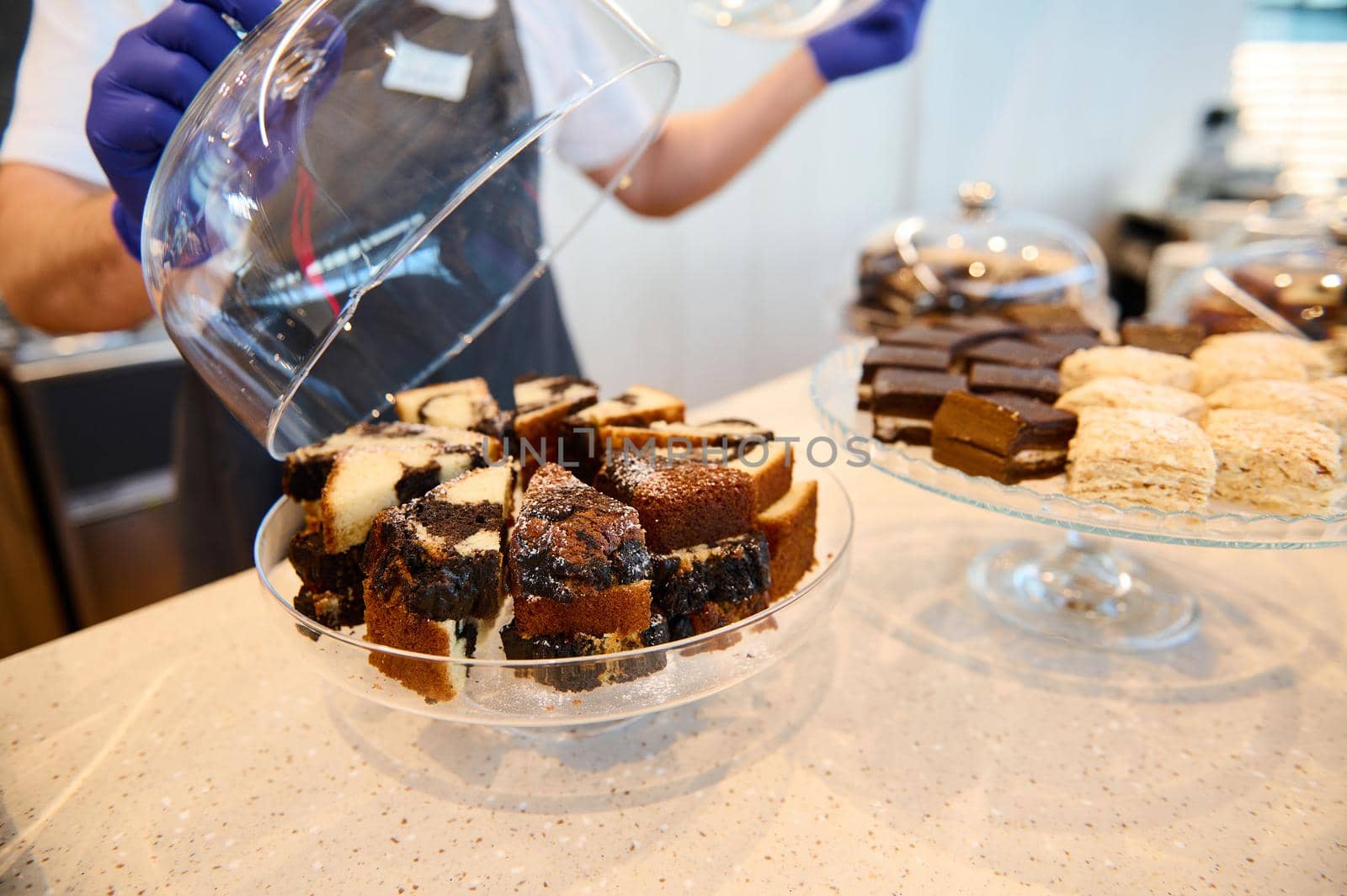 Close-up of chef cook in white uniform and black apron picking up cloche and showing pastries, cakes on the bar table at the open kitchen of the airport restaurant. Image focused on the dessert.