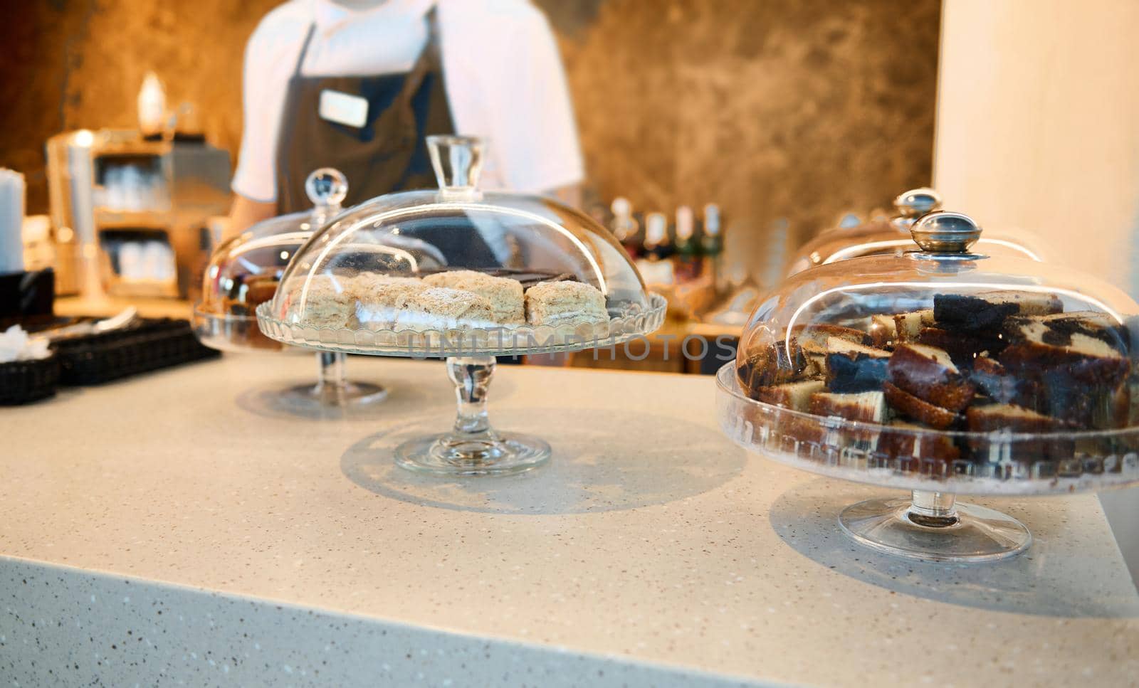 Image focused on cloche with pastries and chocolate cheese creamy desserts on a restaurant countertop with blurred chef in white uniform and black apron on background. Food and drink concept by artgf