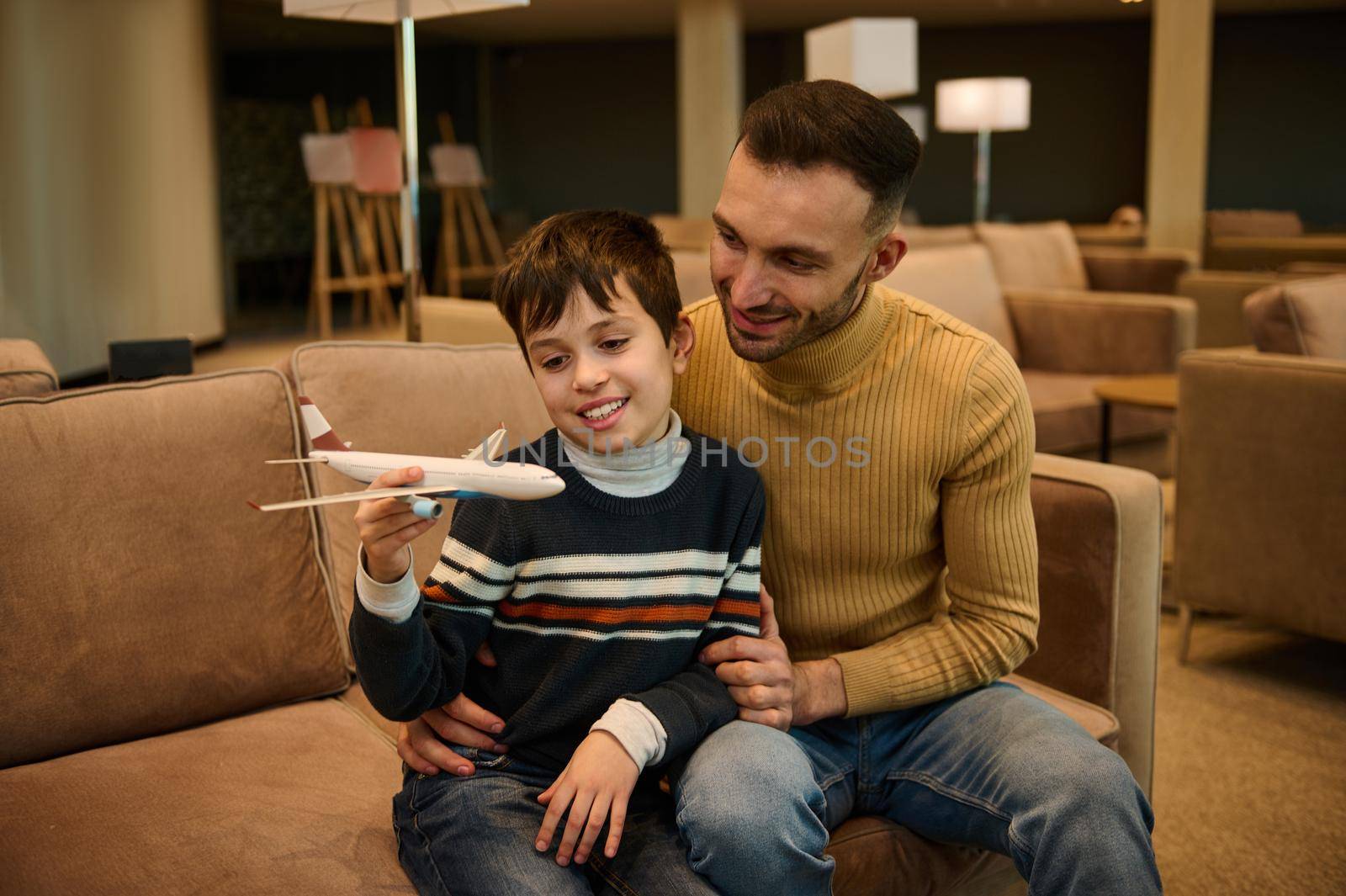 Adorable boy plays with toy plane while his young loving father sits next to him, hugs him while waiting for boarding a flight in the VIP lounge of the departure terminal of the international airport by artgf