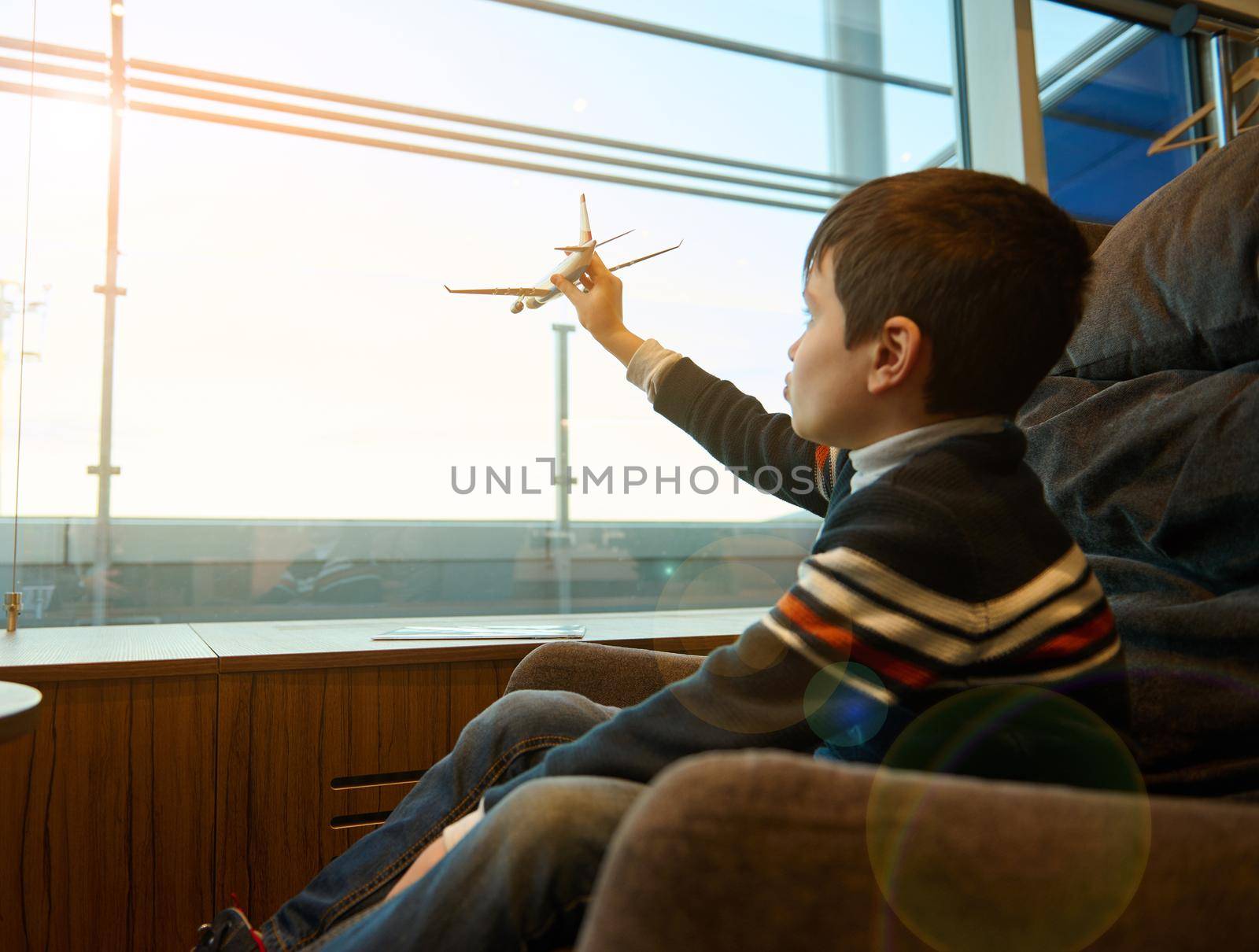 Portrait of adorable child boy holding a toy airplane in outstretched hand imitating flight, against panoramic windows overlooking the runway in the airport departure terminal while waiting for flight by artgf