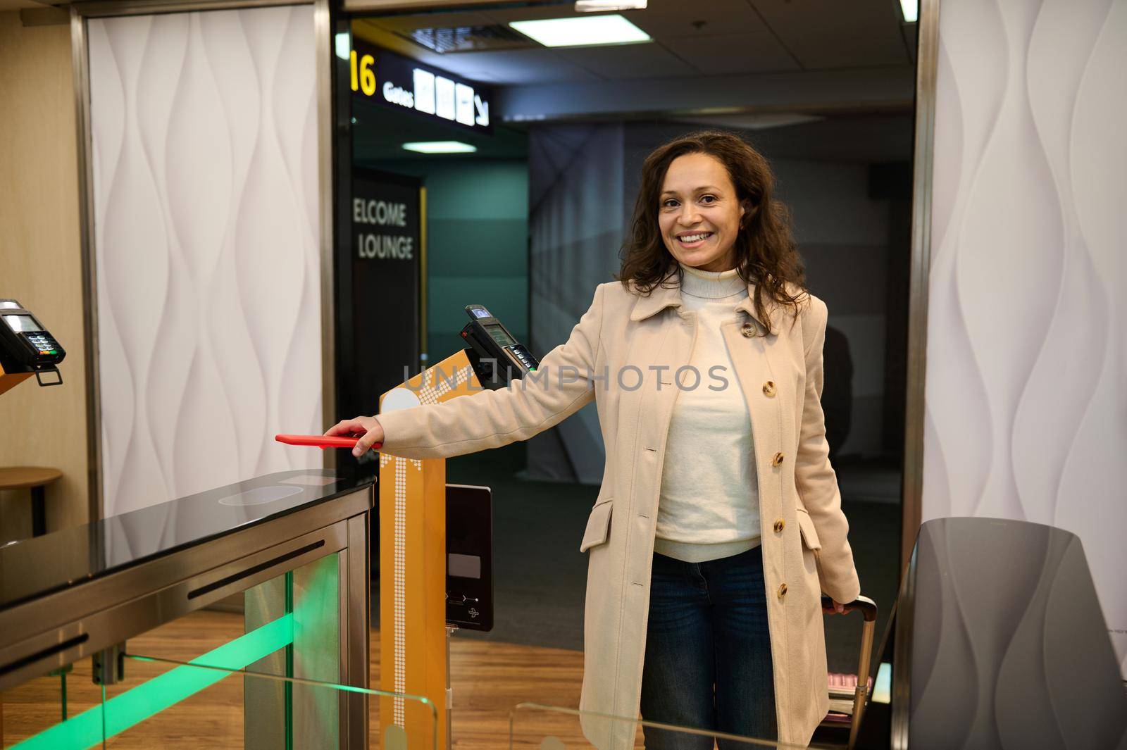 Smiling pretty woman passenger with suitcase scans info on the phone by placing it over the scanner to read the QR-code at the check-in counter while entering VIP lounge in airport departure terminal