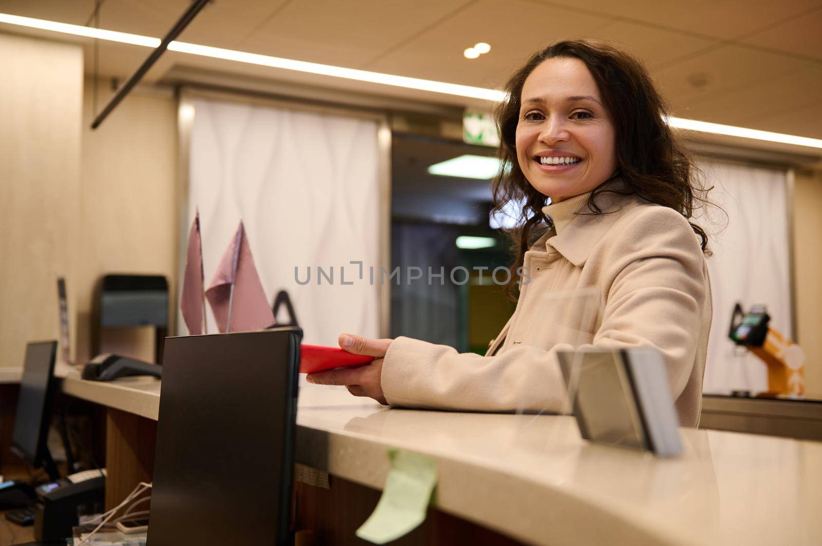 Charming business woman standing with a mobile phone in her hand at the check-in counter in the VIP lounge of the international airport departure terminal, smiling cutely at camera, waiting for flight by artgf