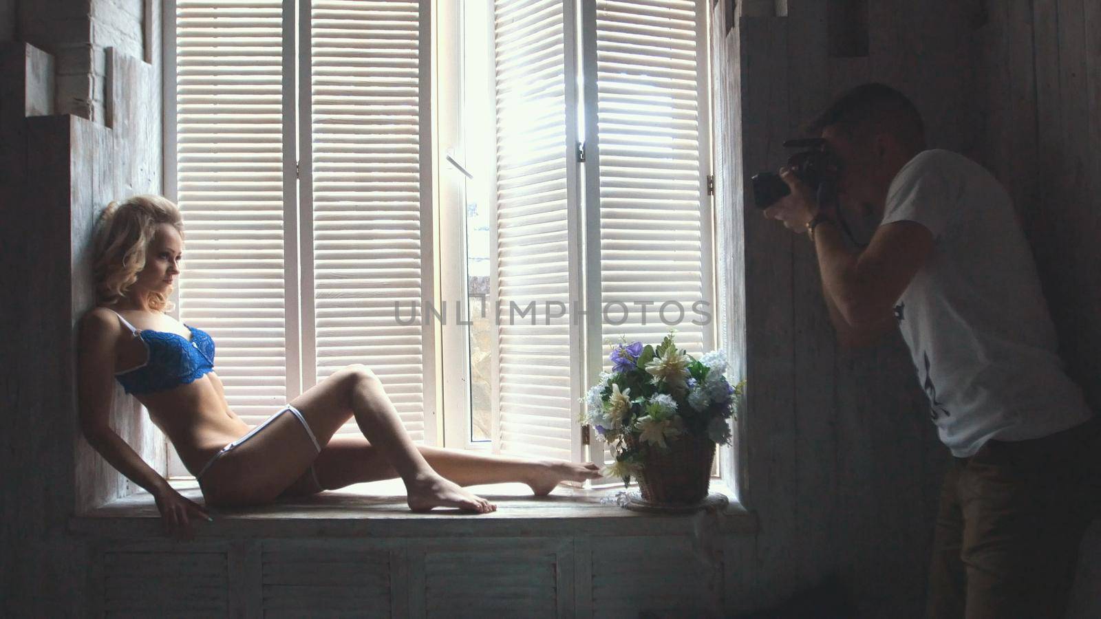 Young blonde model in the blue underwear posing for photographer sitting at the window during fashion photo session, shooting in the studio