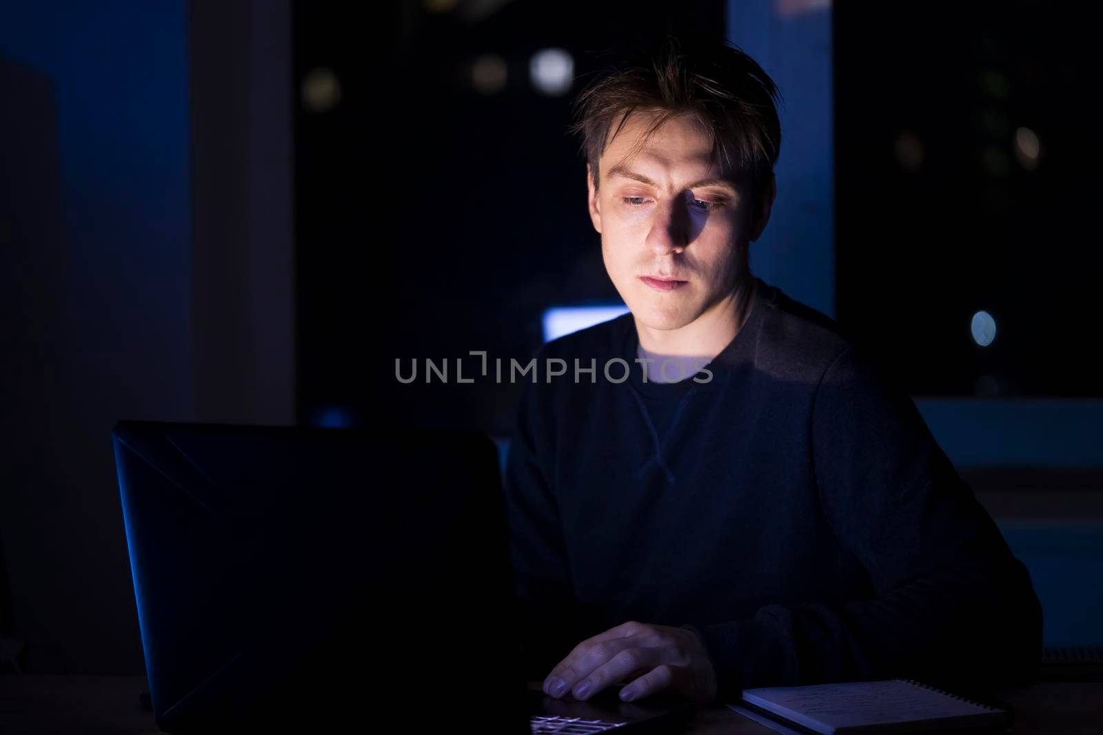 A young man sits and works at night on a laptop on the network, types on the keyboard, writes text messages, studies, prepares a project, a programmer, web designer works as a freelancer at home.