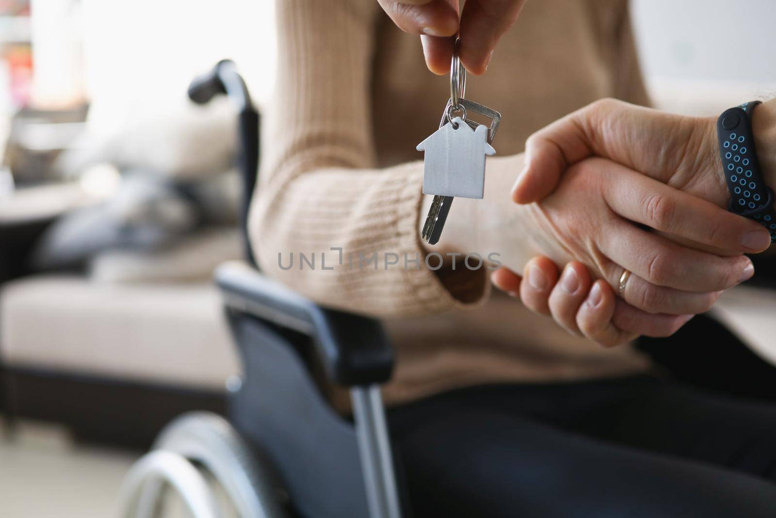 Close-up of realtor give keys from new accommodation to woman in wheelchair, kind handshake in agreement. Disability, government help, real estate concept