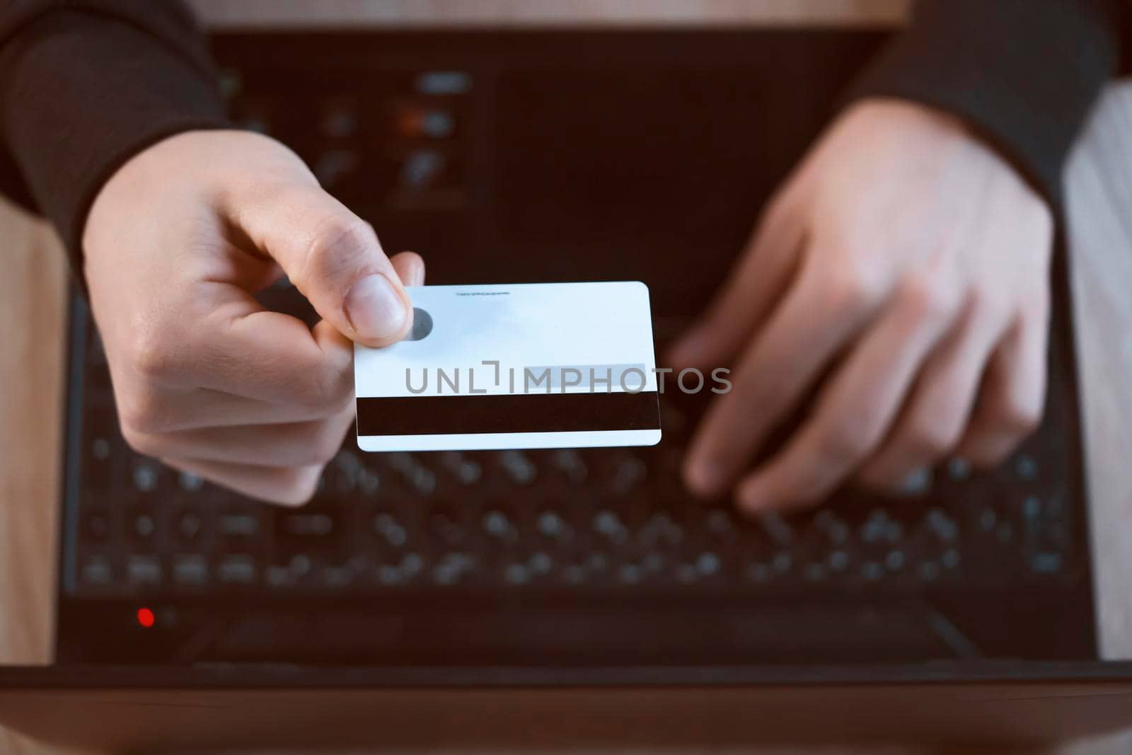 A man holds a bank card in his hand and types on a laptop keyboard, pays for purchases, makes online banking through an application, close-up view from above of his hands.