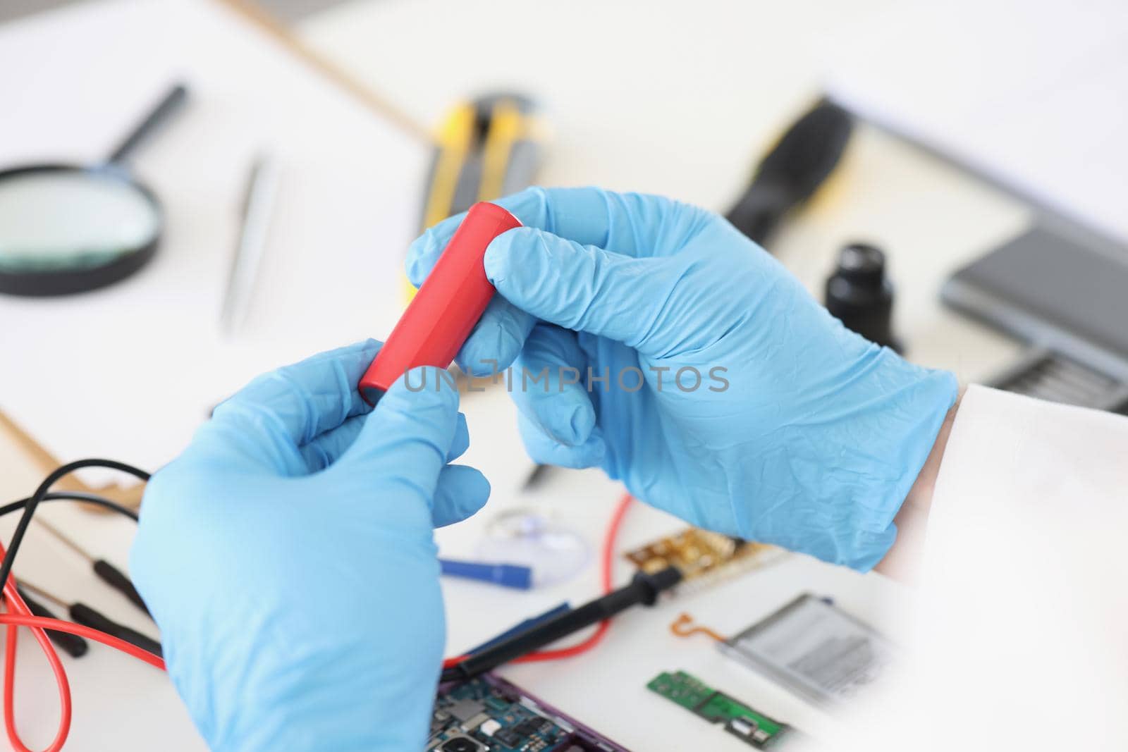 Close-up of male in blue gloves holding red battery piece, repairing broken or damaged device by himself. Workplace full with tools. Handyman, fix concept