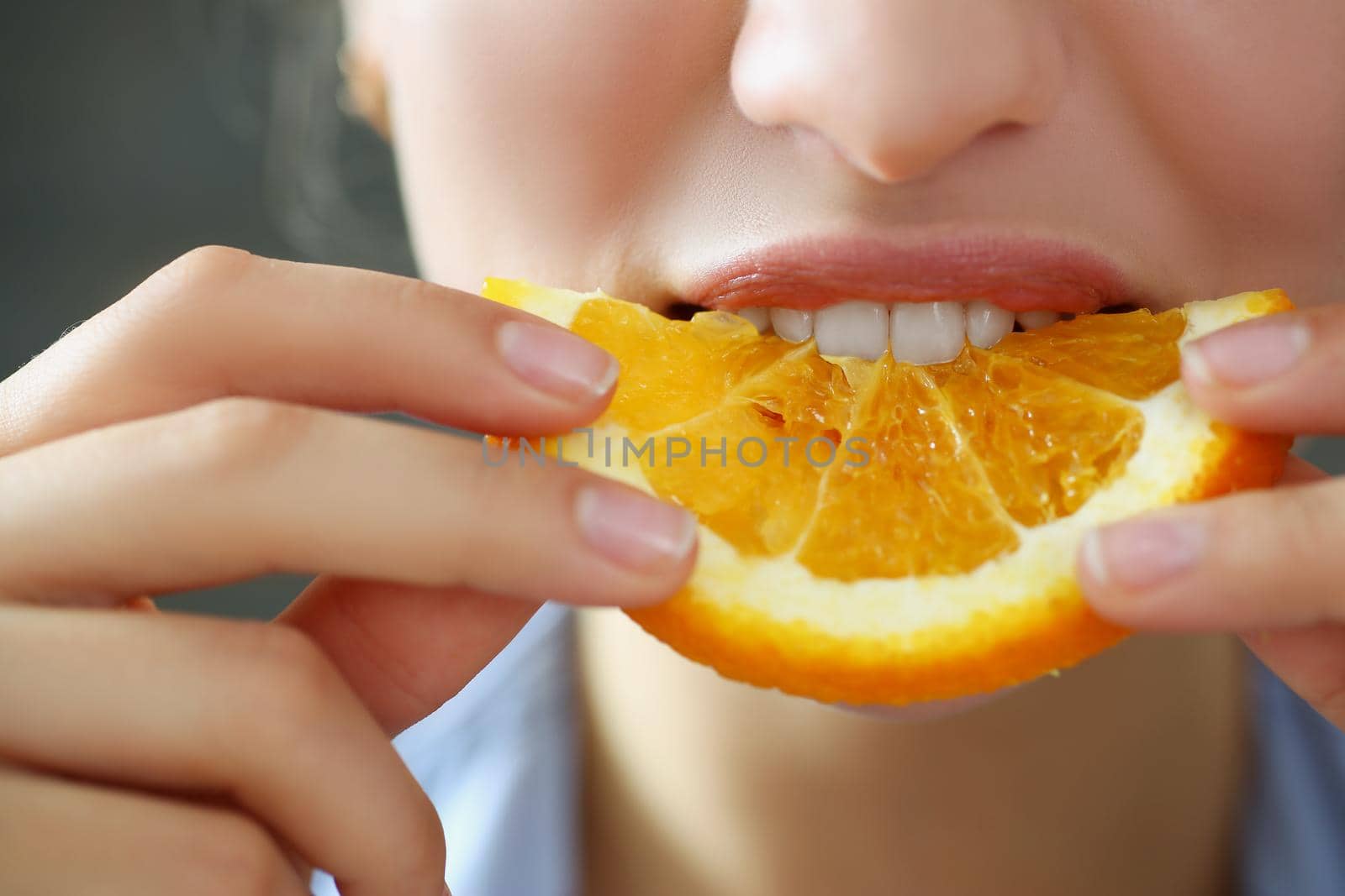 Close-up of woman hold in hand slice of chopped orange eats for breakfast with mouth in kitchen. Healthy eating, diet, juicy, fruit, summer harvest concept