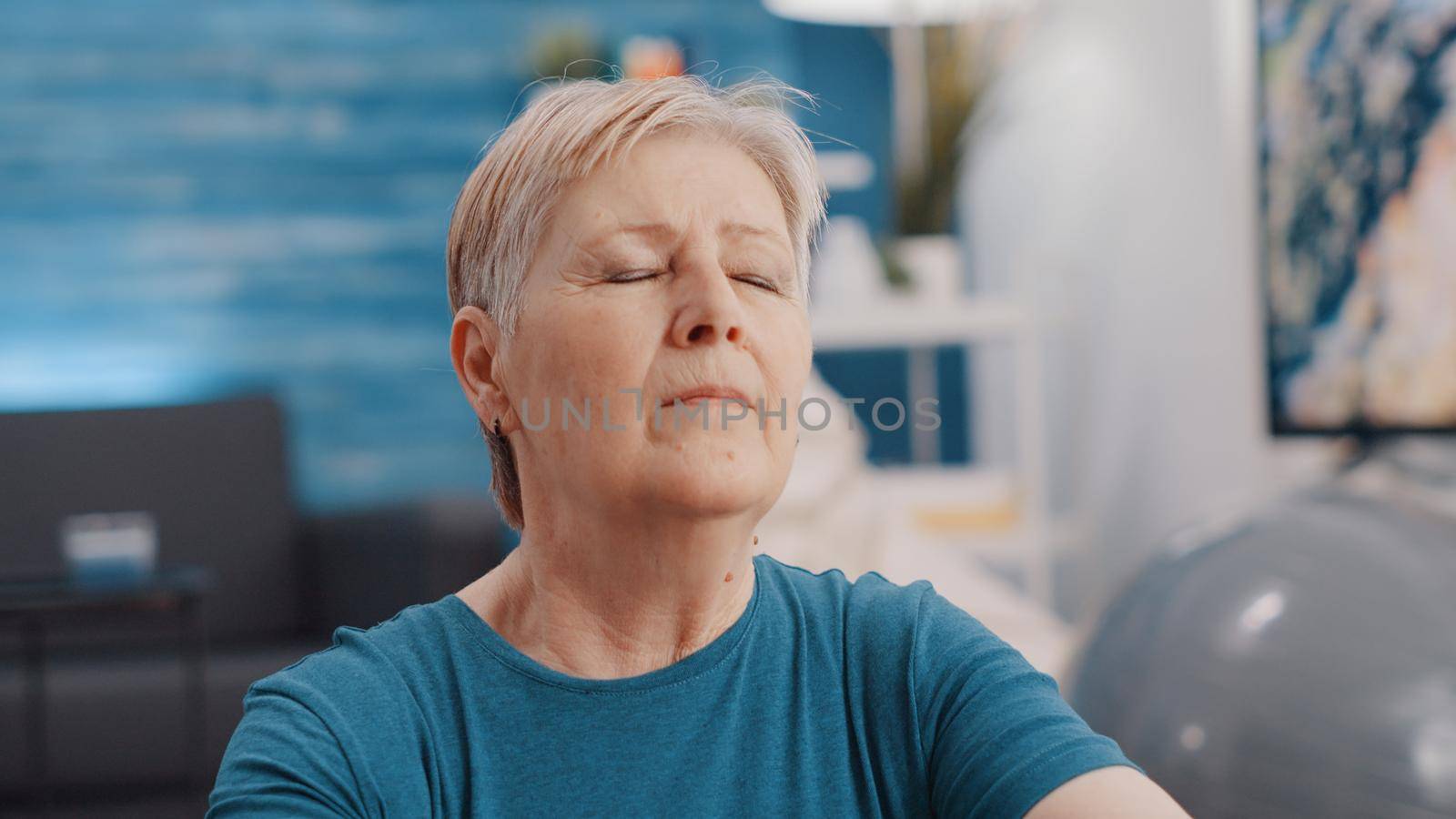 Close up of old woman feeling zen with eyes closed at home. Portrait of senior person relaxing after meditation on yoga mat. Retired adult meditating for wellness and calm recreation