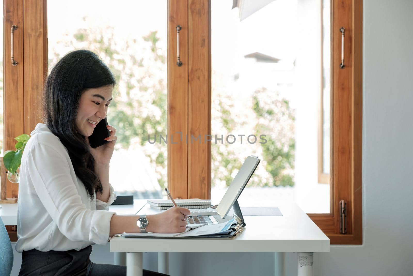 Side view happy asian businesswoman sitting on her workplace in the office. Young woman working for financial document data charts with laptop during call phone in the office.