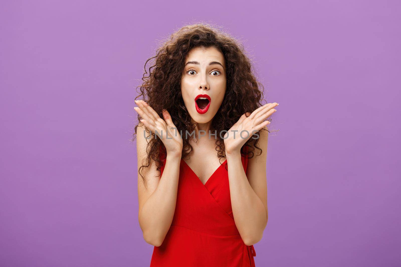 Waist-up shot of excited and amazed expressive elegant adult curly-haired woman with red lipstick in evening luxurious dress gasping from amazement waving palms near face staring surprised at camera by Benzoix