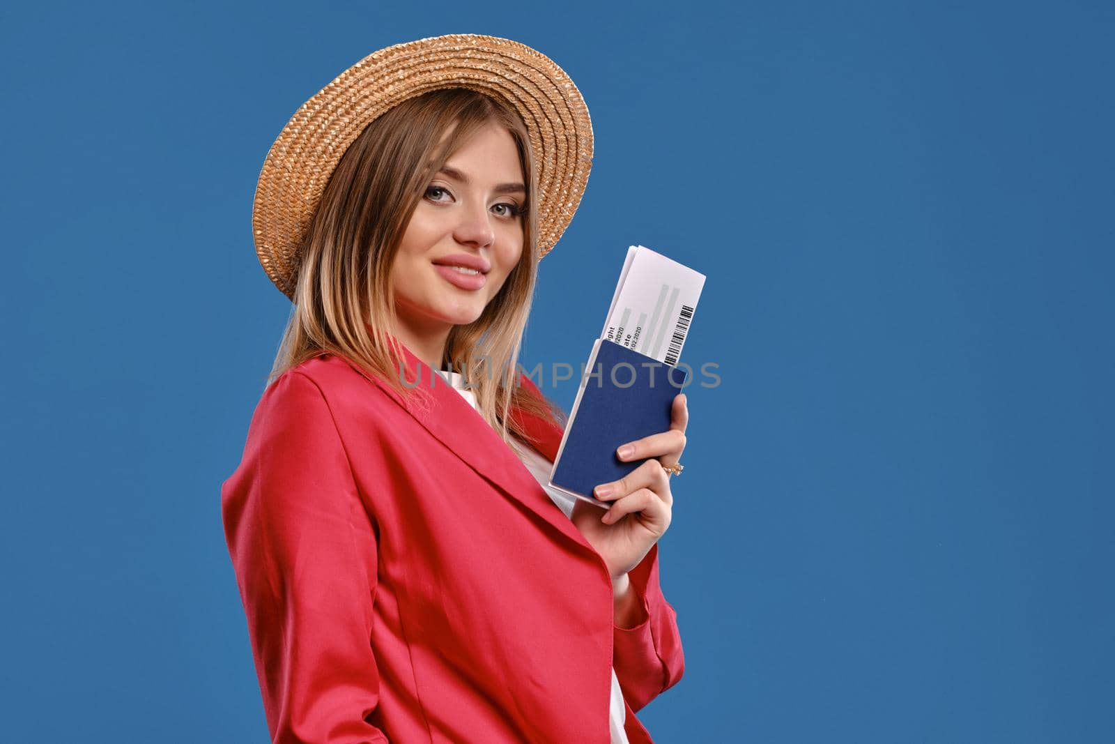 Cute blonde model in straw hat, white blouse and red pantsuit. She is smiling, holding passport and ticket, posing sideways against blue studio background. Travelling concept. Close-up, copy space