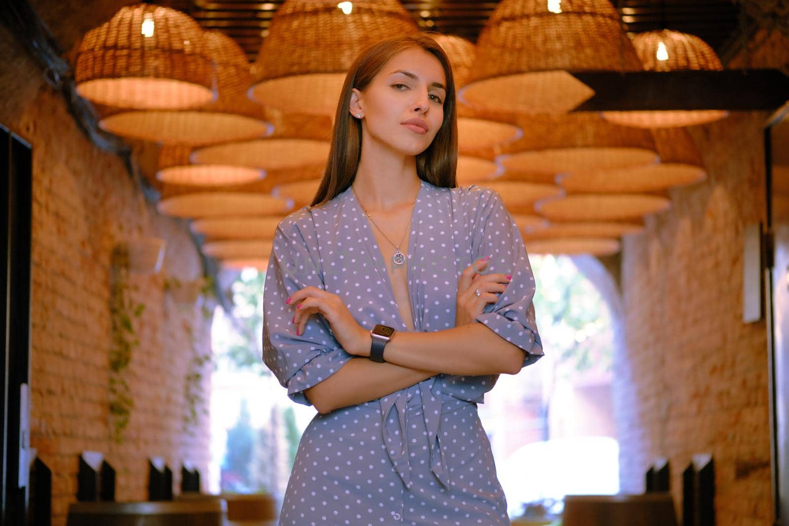 Nice blonde female in a long blue dress with polka-dots, watch, with a pendant around her neck has folded her hands, posing standing under a huge chandeliers in a city cafe while walking alone. The concept of fashion and style. Close-up shot.