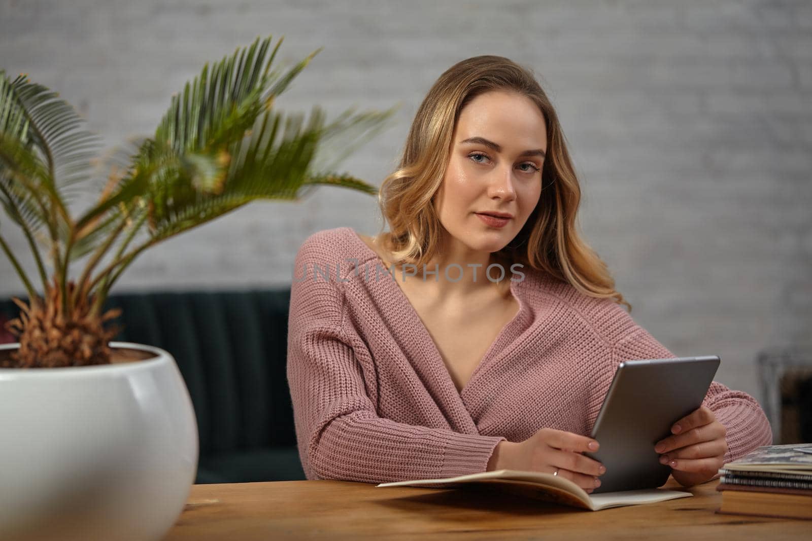 Blonde lady in pink cardigan is holding a tablet. Sitting at wooden table with notebooks and palm tree in pot on it. Student, blogger. Interior with white brick wall and gray sofa. Close-up