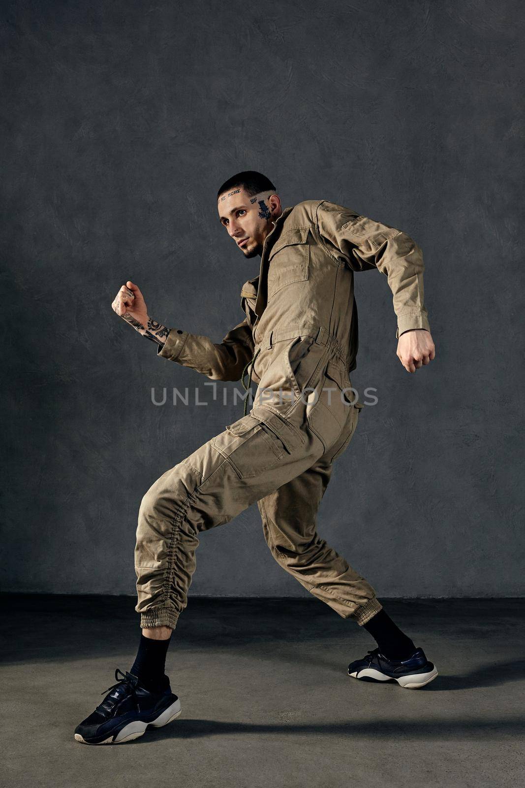 Good-looking man with tattooed body and face, earrings, beard. Dressed in khaki overalls and black sneakers. He is dancing against gray studio background. Dancehall, hip-hop. Full length, copy space