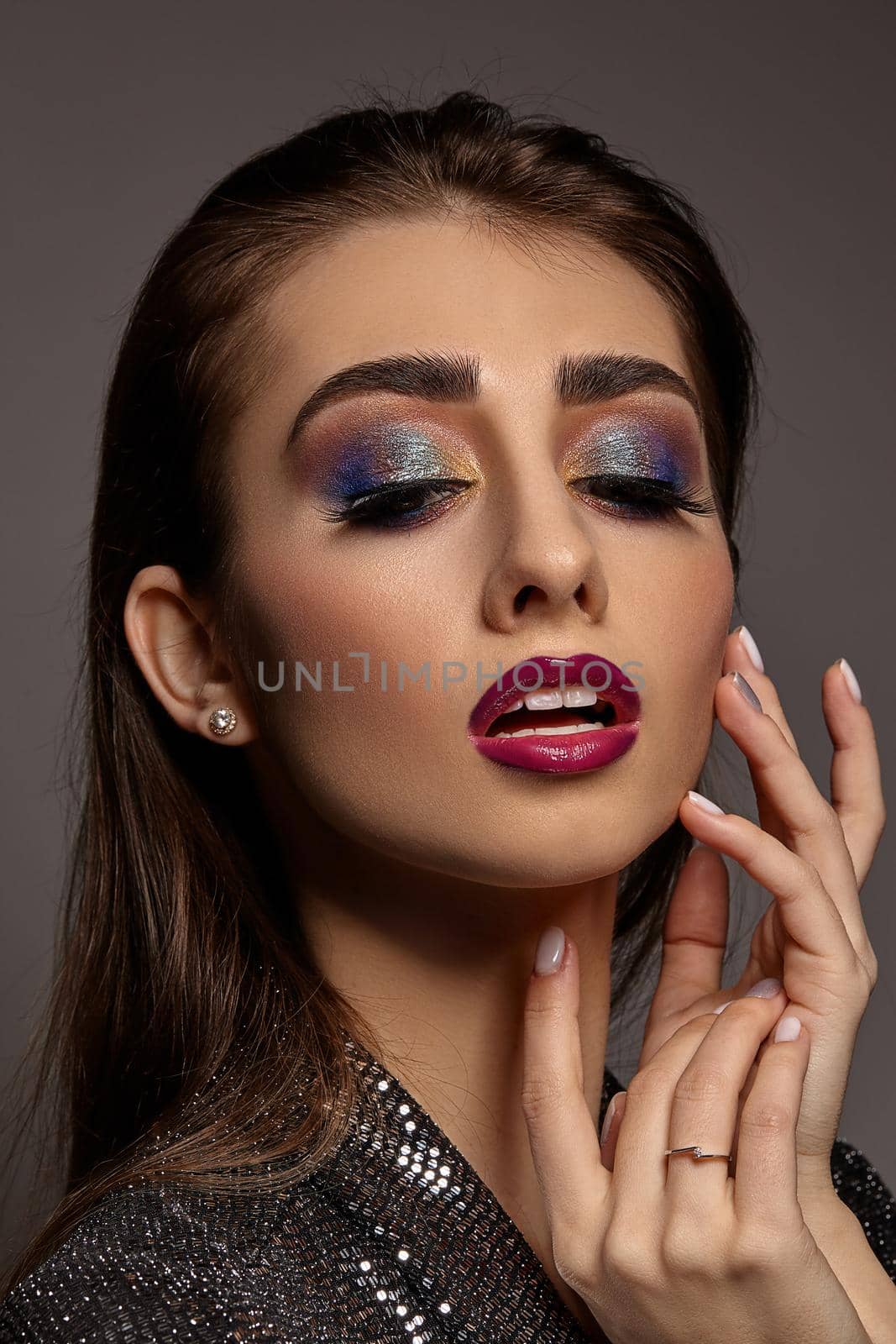 Brunette female in jewelry and black sparkling dress is touching her face, looking down, posing on gray background. Colorful eyeshadow, false eyelashes, glossy red lips. Professional makeup. Close up