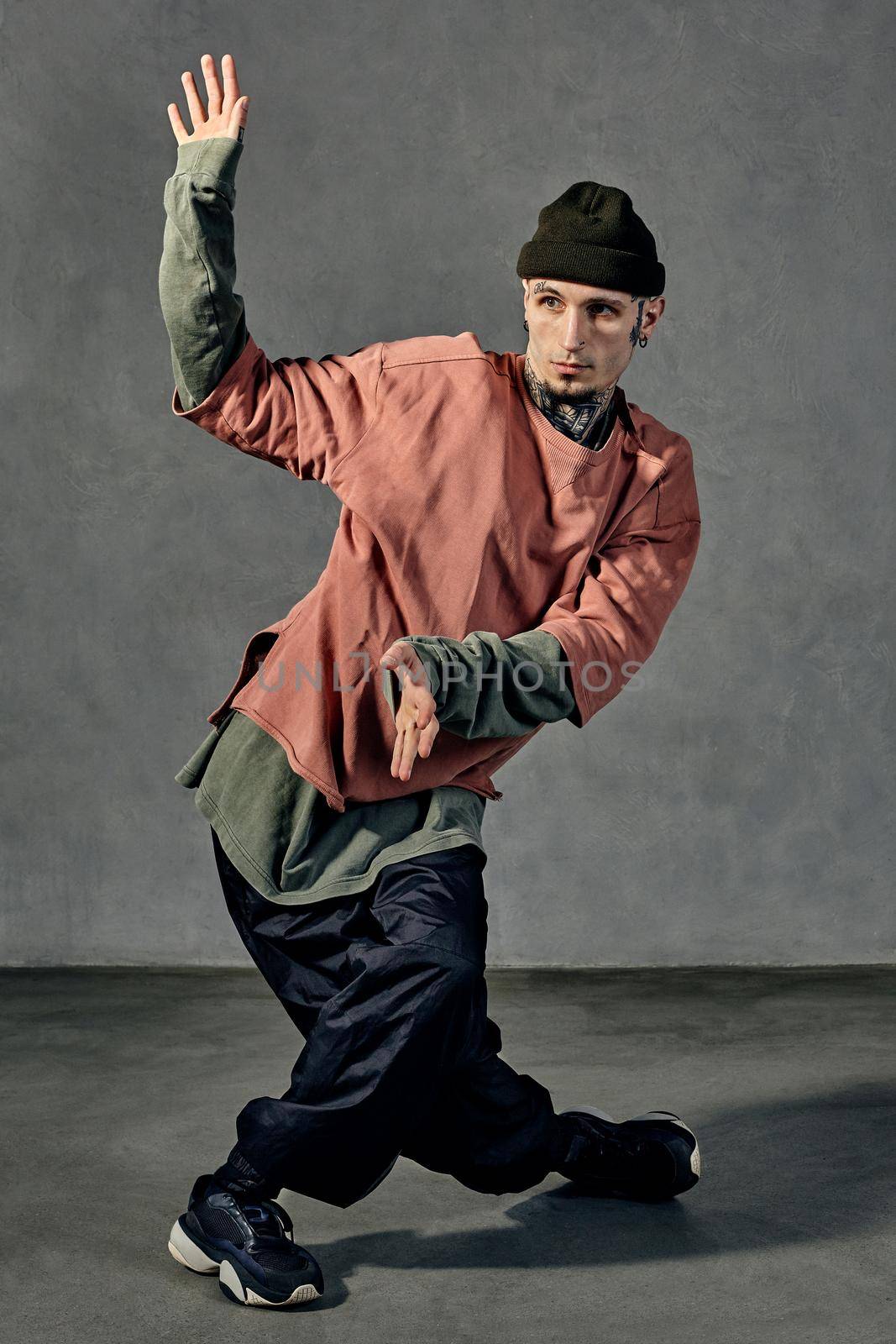 Young graceful man with tattooed body and face, earrings, beard. Dressed in hat, casual clothes and black sneakers. Dancing on gray background. Dancehall, hip-hop. Full length, copy space