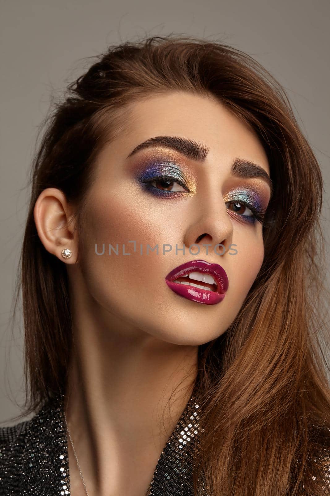 Brunette girl in black shiny dress and jewelry posing on gray background. Luxury makeup, perfect skin. Multi-colored eyeshadow, false eyelashes, glossy burgundy lips. Professional maquillage. Close up