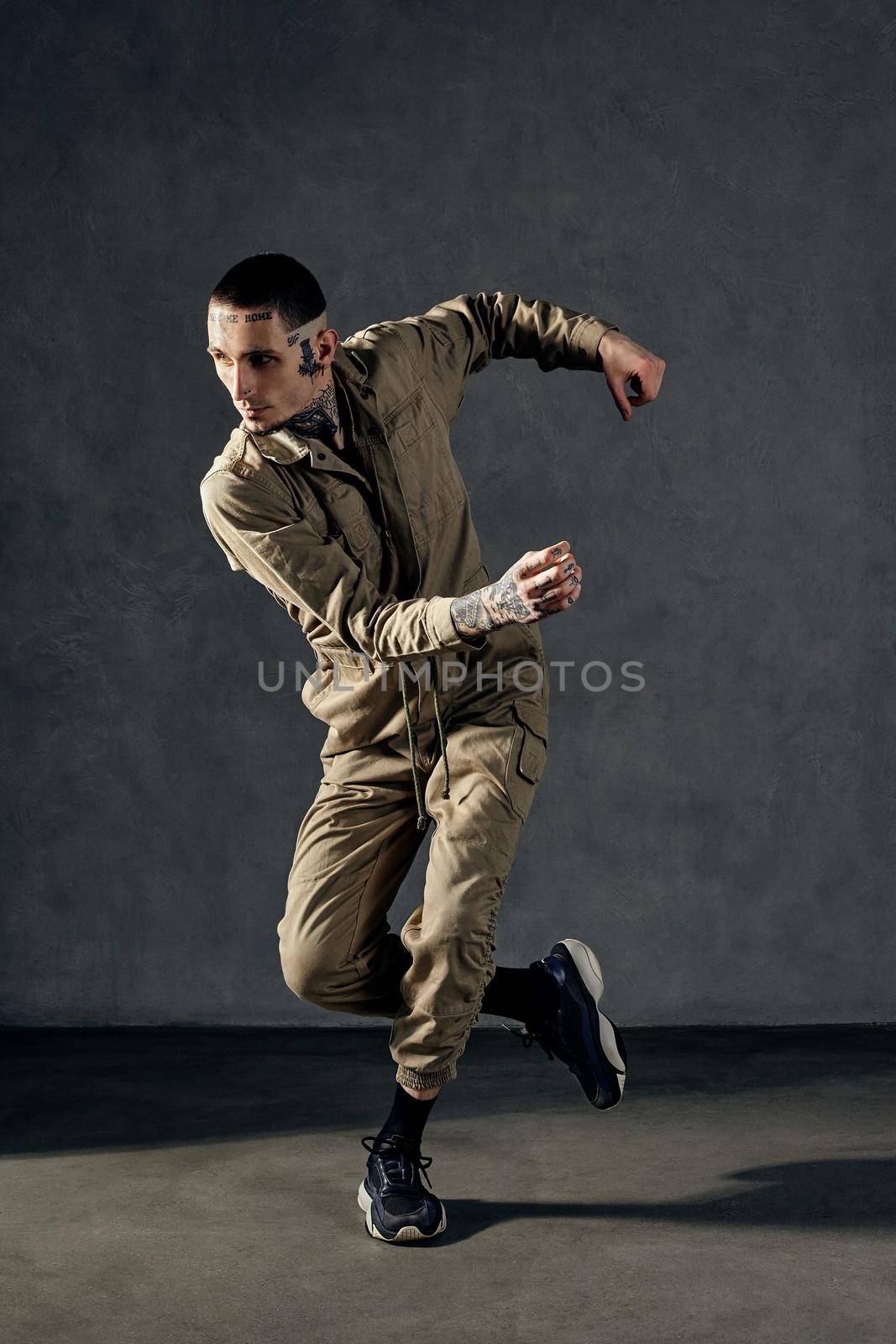 Young modern guy with tattooed body and face, earrings, beard. Dressed in khaki overalls and black sneakers. He is dancing against gray studio background. Dancehall, hip-hop. Full length, copy space