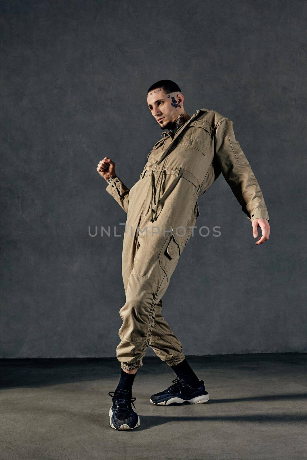 Young flexible fellow with tattooed body and face, earrings, beard. Dressed in khaki jumpsuit and black sneakers. He dancing against gray studio background. Dancehall, hip-hop. Full length, copy space