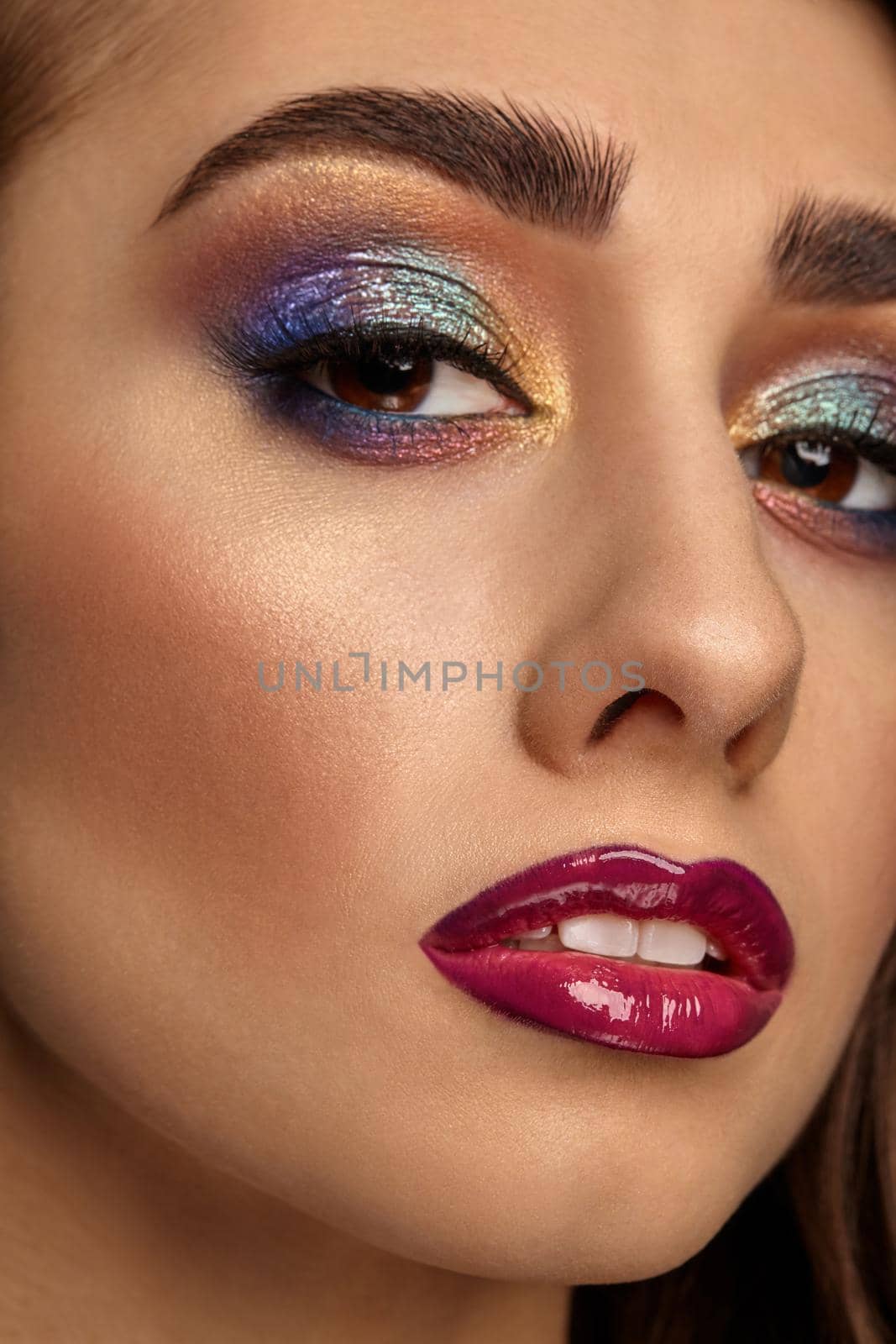 Brunette female with luxury makeup and perfect skin is looking at you. Multi-colored eyeshadow, false eyelashes, glossy burgundy lips. Professional maquillage. Close up