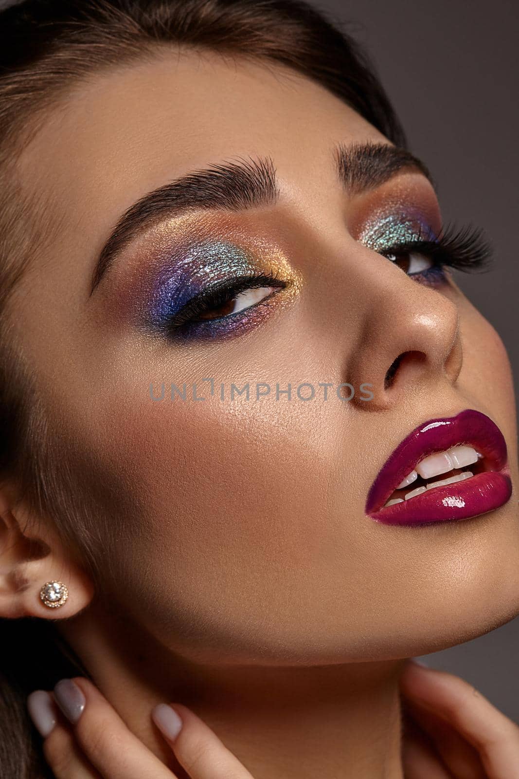 Brunette girl is touching her neck, posing sideways on gray background. Colorful eyeshadow, false eyelashes, glossy purple lips and white teeth. Luxury makeup. Professional maquillage. Close up