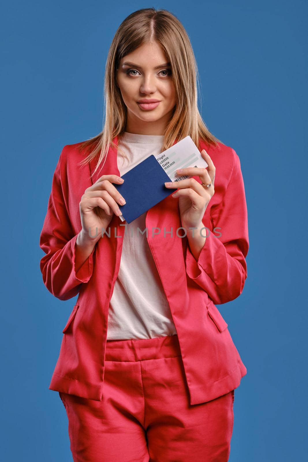 Blonde woman in white blouse and red pantsuit. She smiling, holding passport and ticket while posing on blue studio background. Close-up by nazarovsergey