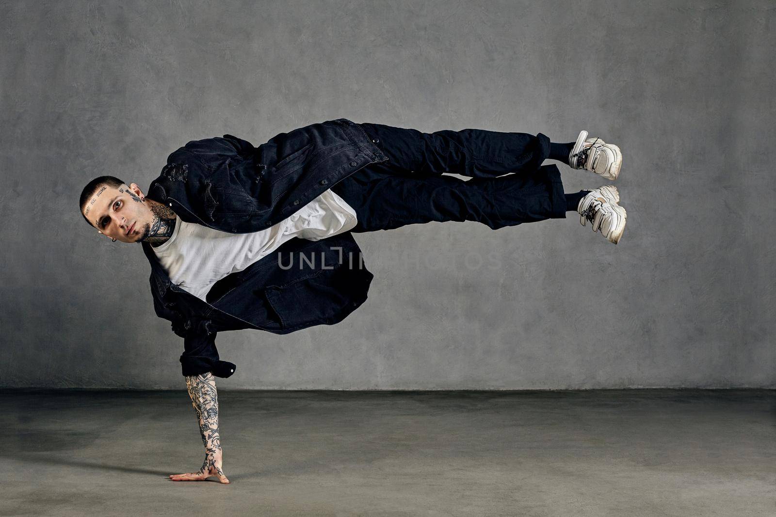 Flexible fellow with tattooed body and face, earrings, beard. Dressed in white t-shirt and sneakers, black denim shirt, pants. Performing tricks on gray background. Dancehall, hip-hop. Full length