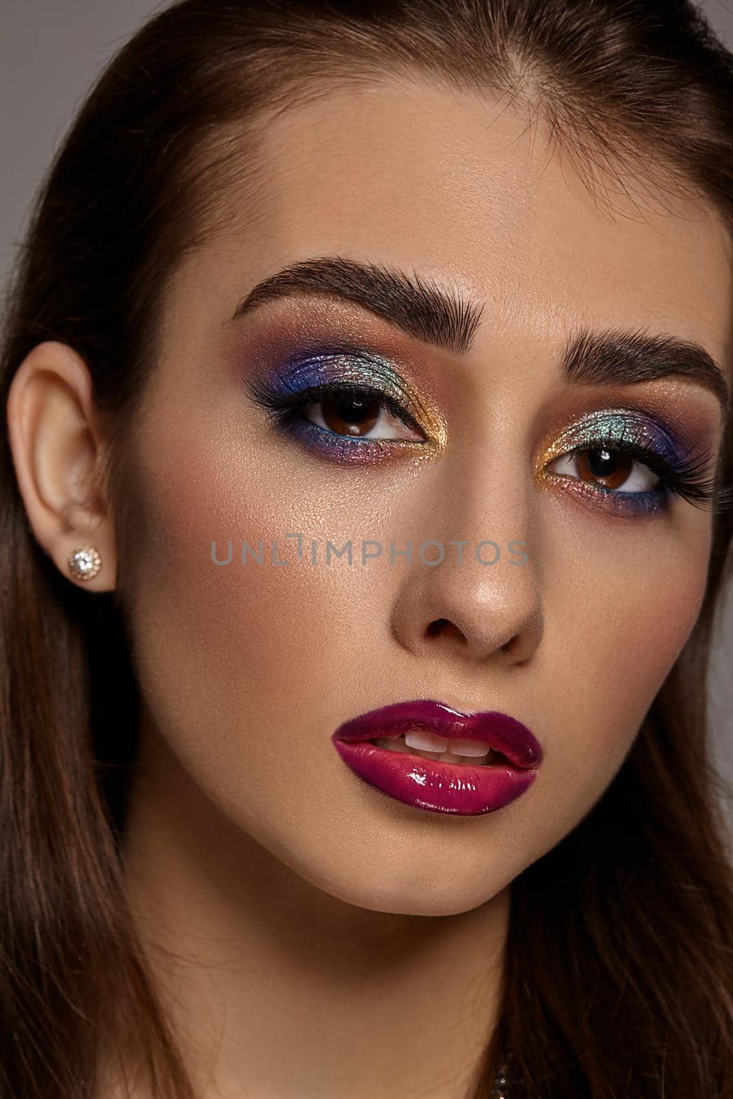 Brunette girl in earrings looking at you while posing on gray background. Luxury makeup, perfect skin. Multi-colored eyeshadow, false eyelashes, glossy purple lips. Professional maquillage. Close up