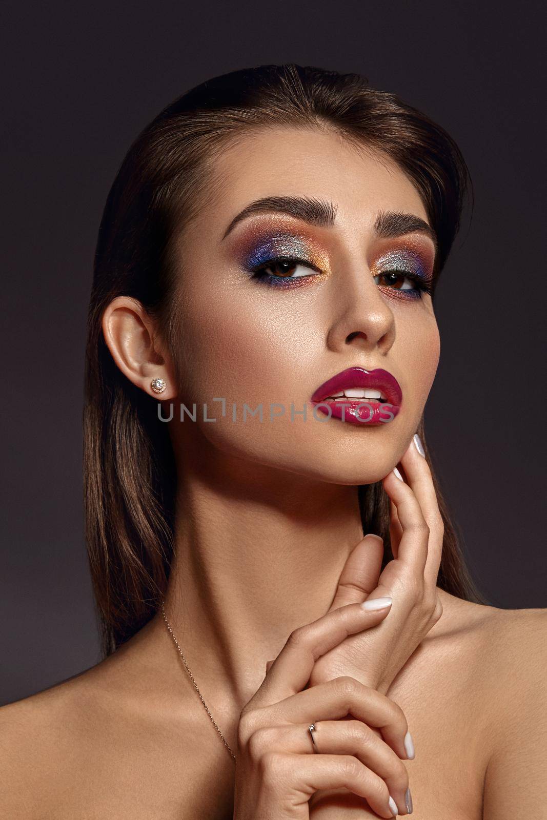 Brunette naked lady in jewelry is touching her face, posing against gray studio background. Luxury makeup, perfect skin. Colorful eyeshadow, false eyelashes, glossy red lips and white teeth. Close up