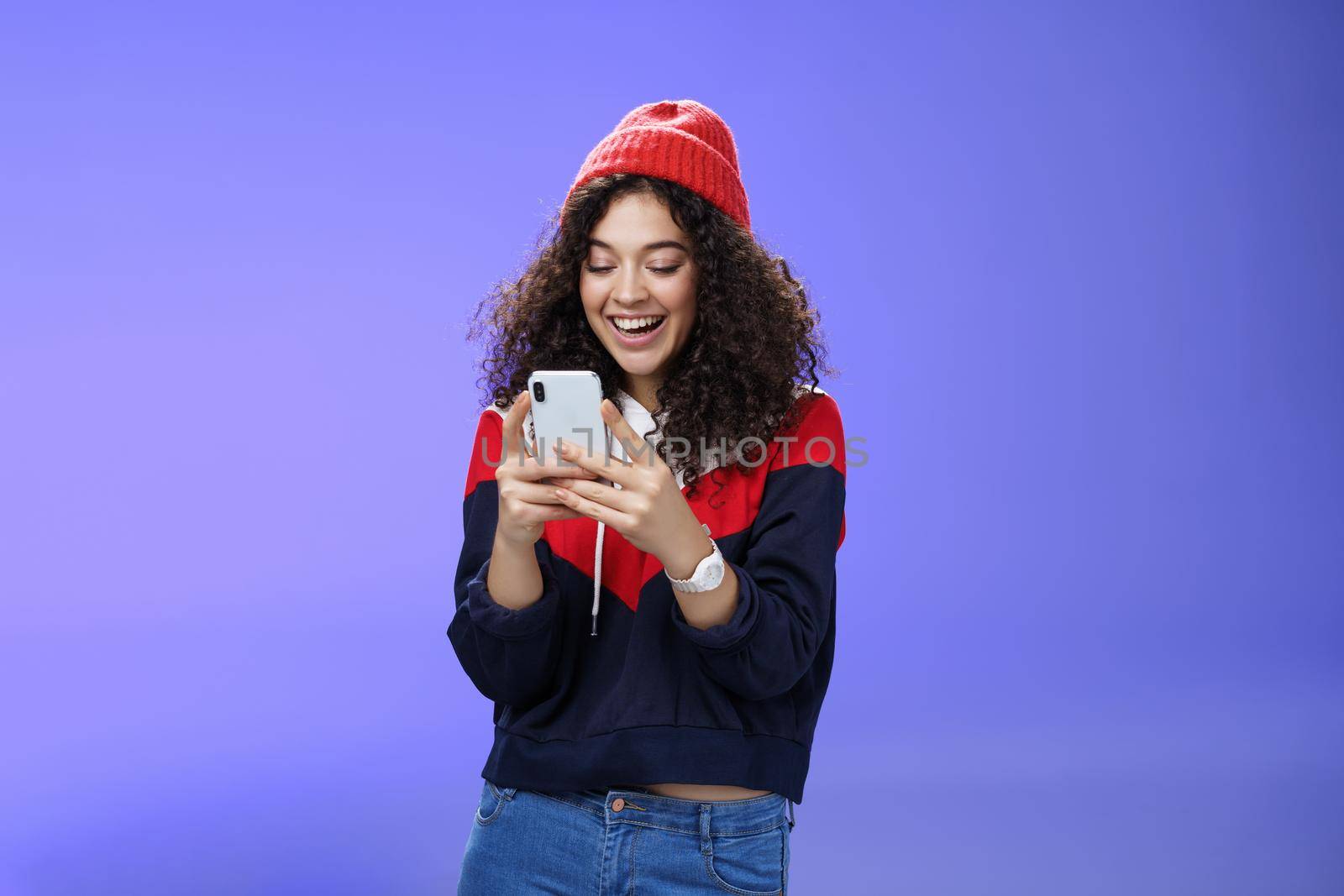 Joyful and sociable stylish caucasian woman with curls in warm beanie having fun laughing and chuckling as using smartphone typing funny message looking at mobile phone screen happily over blue wall by Benzoix