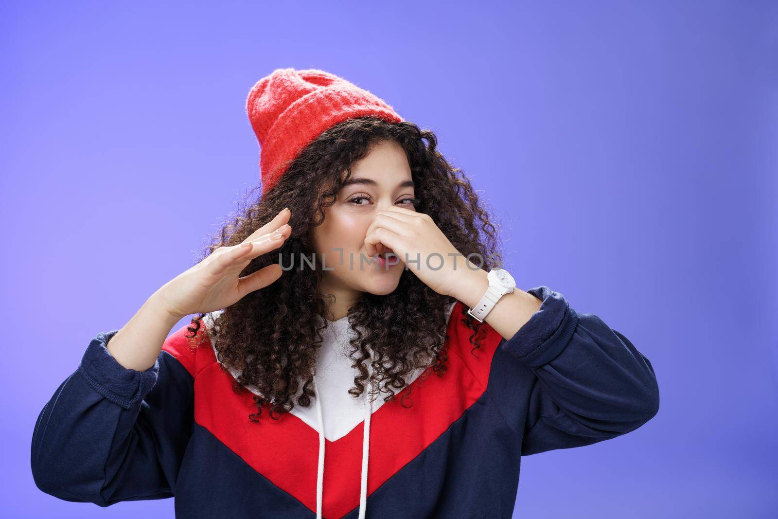 Gosh what stink. Portrait of cute caucasian girl with curly hair in cute trendy hat close nose with fingers and waving hand as smelling awful reek or smell standing disgusted against blue background.