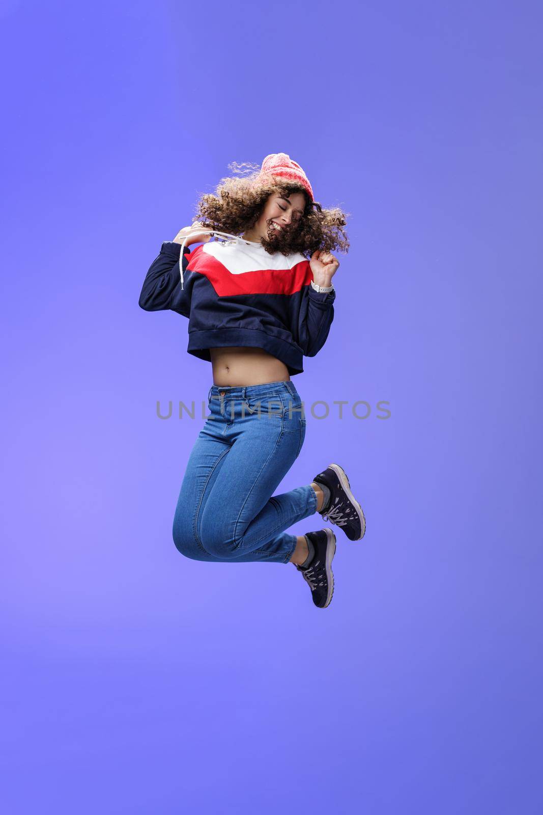 Girl jumping from happiness and delight feeling carefree close eyes and smiling broadly enjoying moment having fun raising hands being dreamy and playful over blue background. Lifestyle, fashion and positive emotions concept